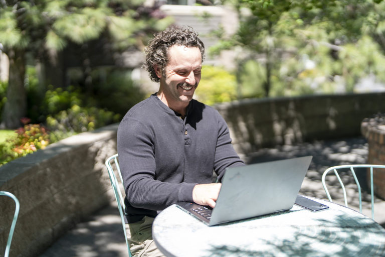 A confident man at an outdoor table using a laptop with a smile on his face.