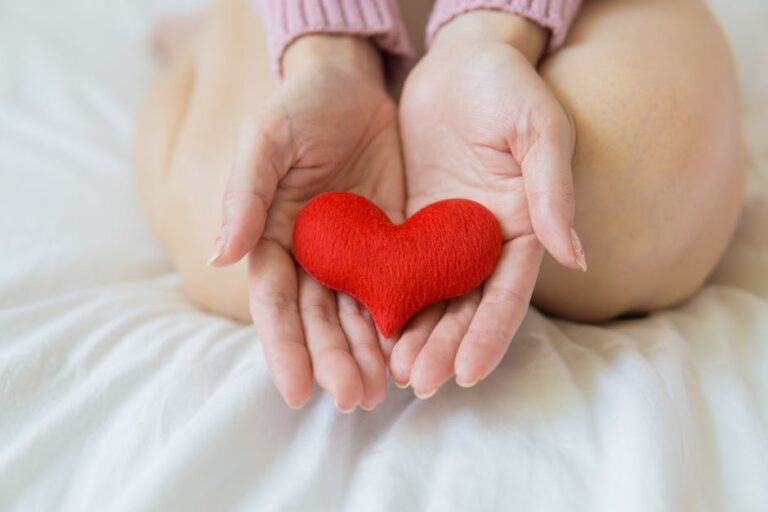 A woman's hands holding a red heart on a bed.