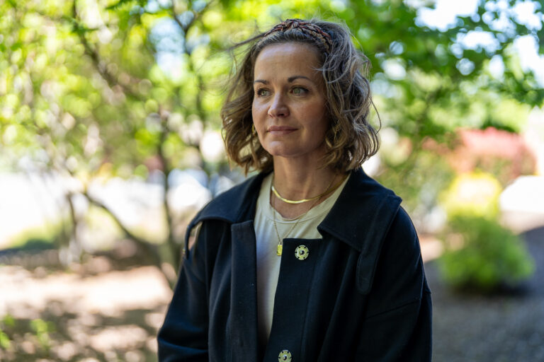 A woman with wavy brown hair wearing a black coat and gold necklace stands outdoors in front of green foliage.
