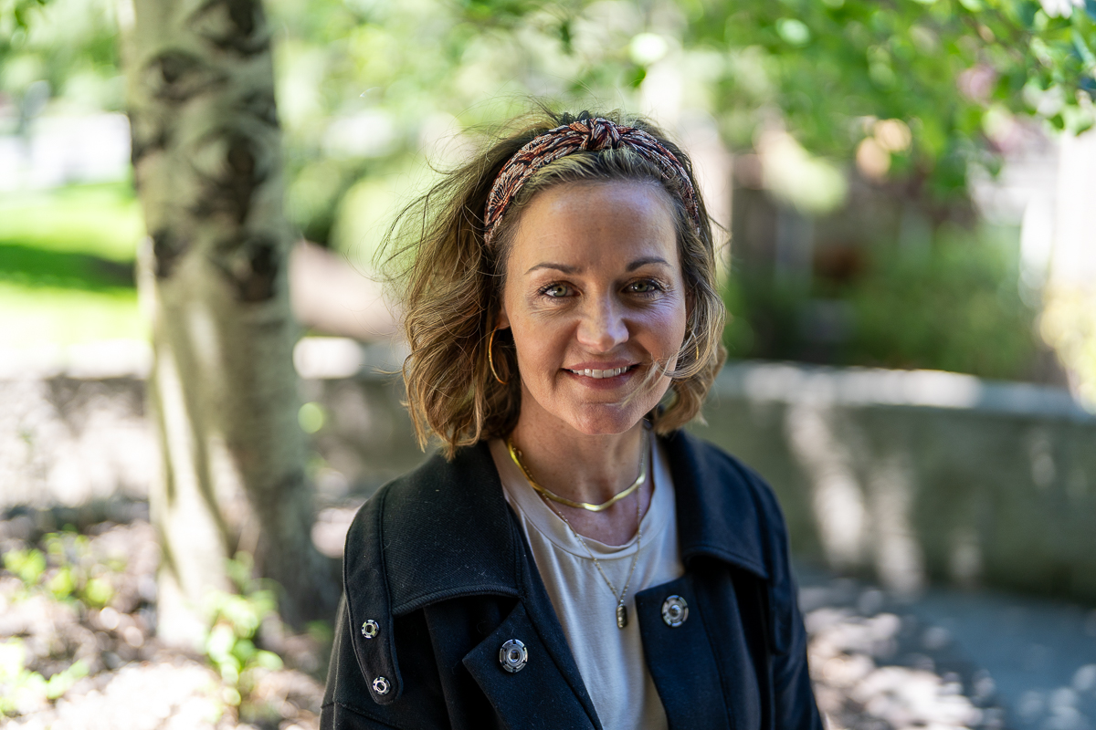 A woman with shoulder-length hair and a headband stands outdoors in front of a tree, smiling at the camera on a sunny day.