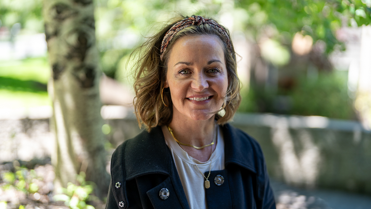 A woman with shoulder-length hair and a headband smiles outdoors, wearing a dark jacket and gold hoop earrings, with trees and sunlight in the background.