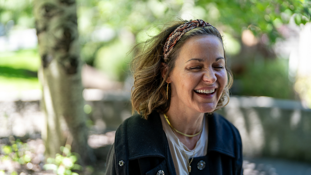 A woman with shoulder-length hair and a headband is smiling with her eyes closed outdoors in a sunlit, leafy setting.
