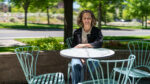 A woman sits alone at a round metal table with empty chairs in an outdoor setting on a sunny day, with greenery and a road in the background.