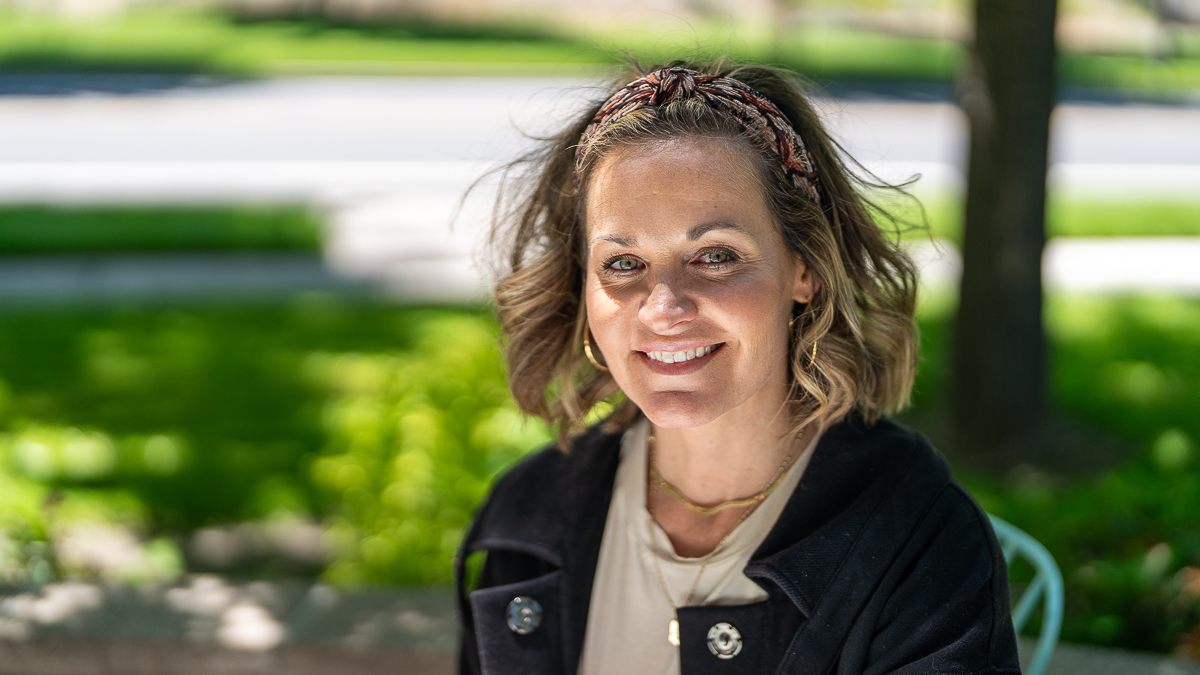 A woman with wavy hair and a headband sits outdoors, smiling slightly. She is wearing a black jacket and light-colored top, with greenery and a road in the blurred background.