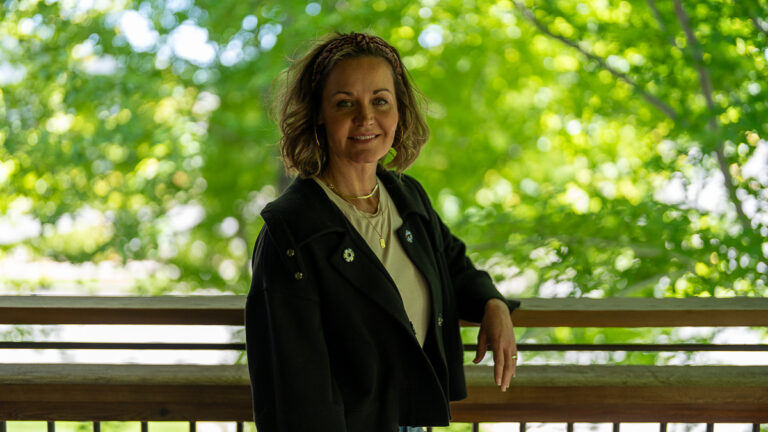 A woman with curly brown hair, wearing a black jacket and light shirt, stands outdoors by a wooden railing with green trees in the background.