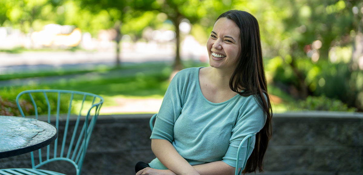 A woman with long dark hair sits outdoors on a metal chair, smiling, with greenery and a stone wall in the background.