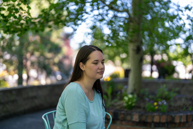A woman with long brown hair sits on a light blue chair outdoors, surrounded by trees and greenery, with a brick planter in the background.