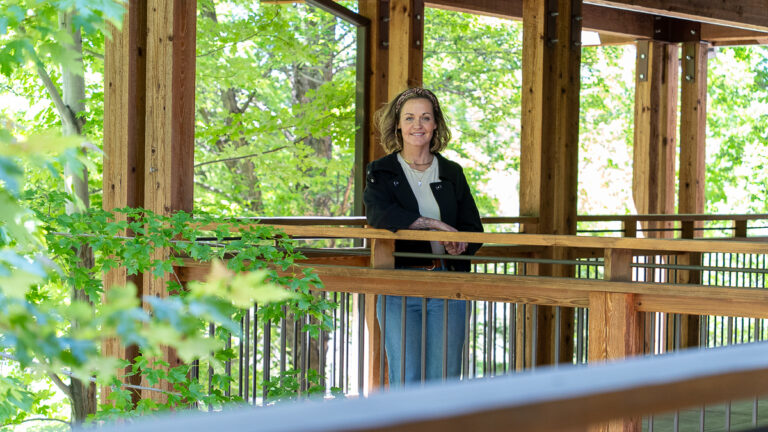 A woman stands and smiles on a wooden outdoor walkway surrounded by green trees and sunlight.