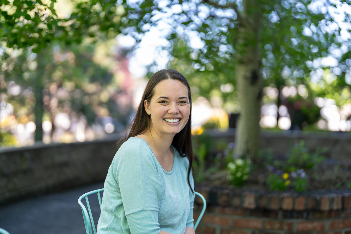 A woman with long brown hair, wearing a light blue top, sits and smiles outdoors in a garden setting with trees and a brick planter in the background.