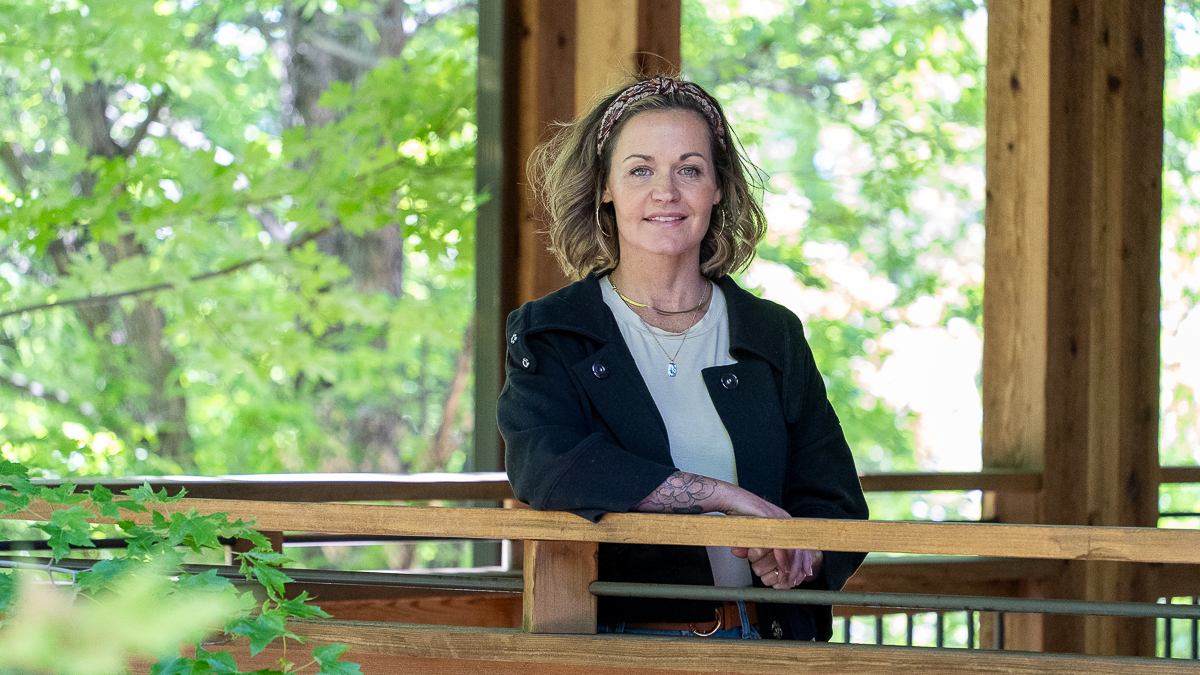 A woman with shoulder-length hair stands at a wooden railing in an outdoor structure surrounded by green trees, wearing a dark jacket over a light shirt.
