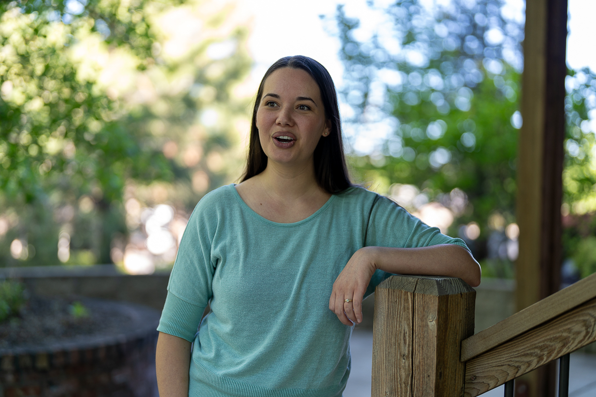 A woman with long dark hair, wearing a light blue top, stands outdoors leaning on a wooden railing, with greenery and trees in the background.