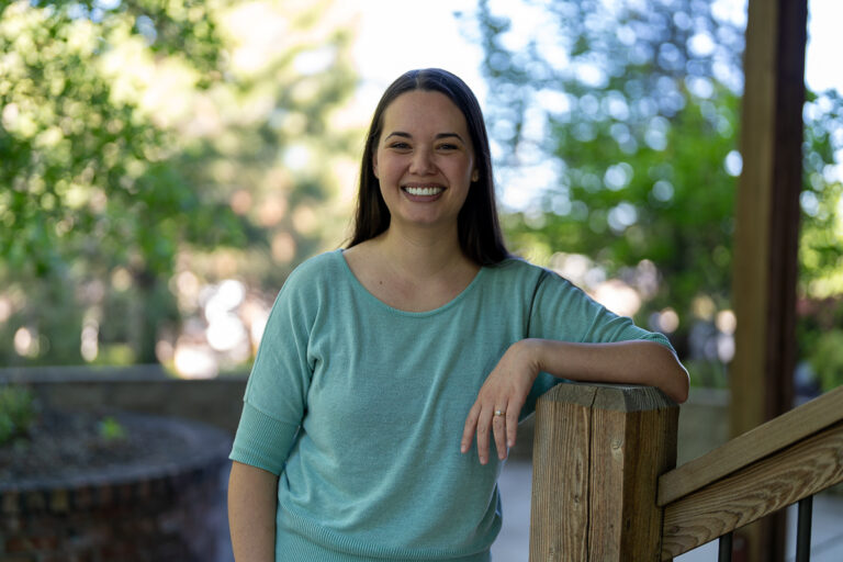 A woman with long dark hair, wearing a light teal top, stands outdoors by a wooden railing, smiling at the camera with greenery in the background.