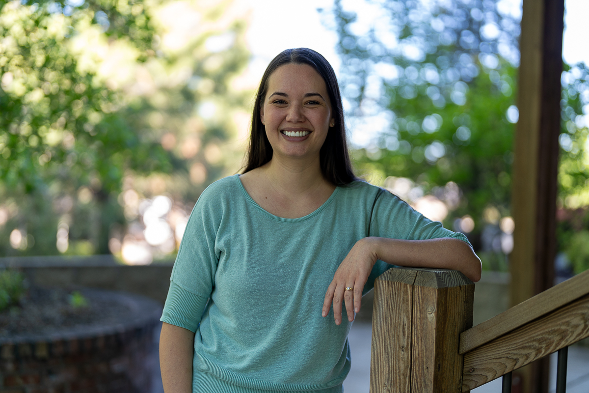 A woman with long dark hair, wearing a light teal top, stands outdoors by a wooden railing, smiling at the camera with greenery in the background.
