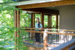 A woman stands and smiles on a wooden balcony surrounded by green trees on a sunny day.