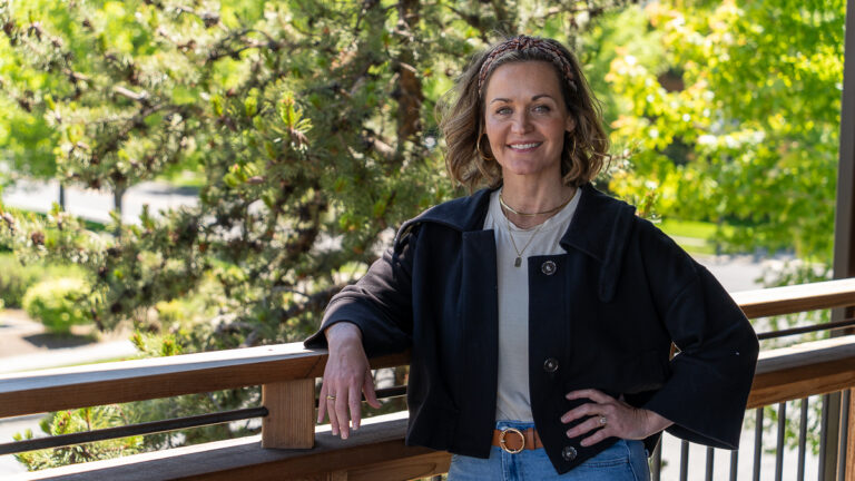 A woman stands outdoors on a wooden balcony, smiling with one hand on her hip and the other resting on the railing. Green trees and sunlight are visible in the background.