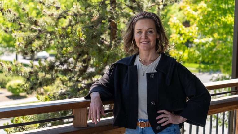 A woman stands outdoors on a wooden balcony, smiling, with trees and greenery in the background.