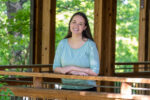 A woman with long brown hair, wearing a light blue top, stands and smiles by a wooden railing in a covered outdoor area with green trees in the background.