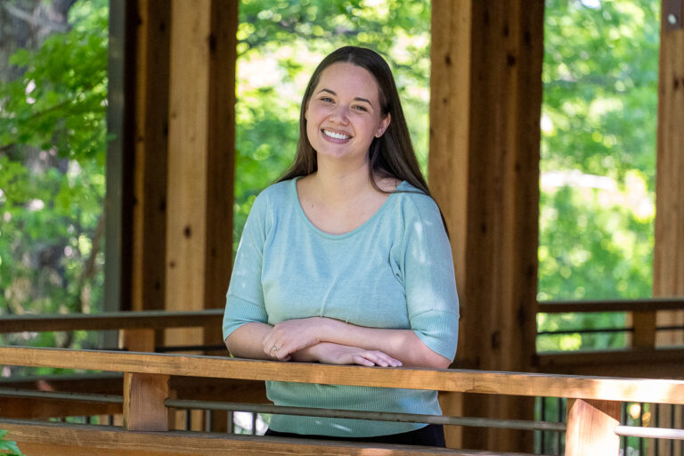 A woman with long brown hair, wearing a light blue top, stands and smiles while leaning on a wooden railing in an outdoor covered area with greenery in the background.