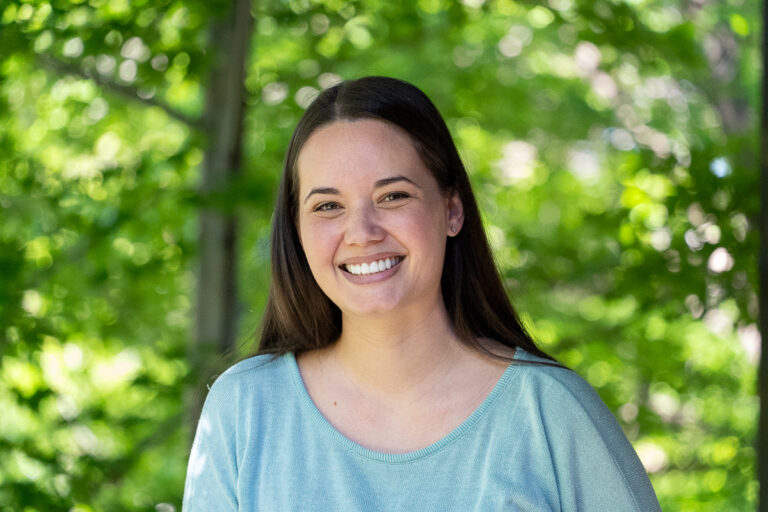 A woman with long brown hair and a light blue shirt smiles outdoors with green foliage in the background.