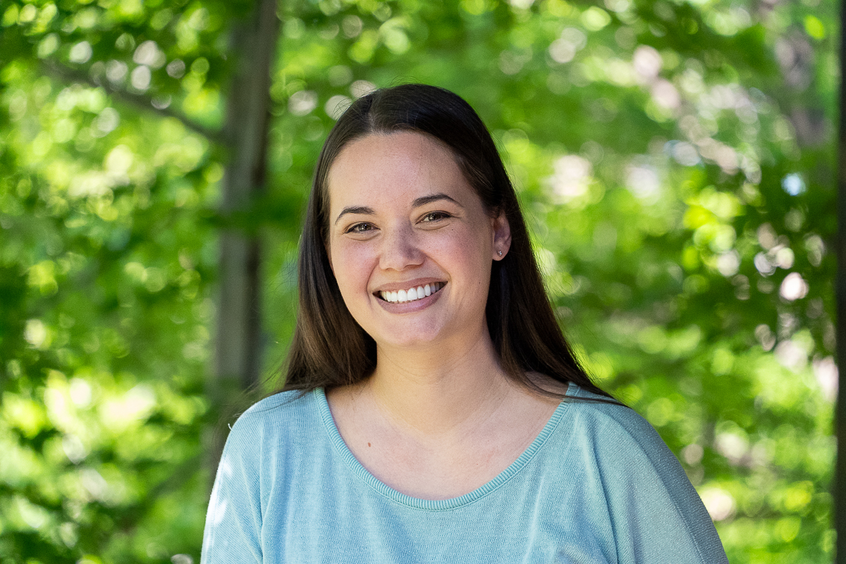 A woman with long brown hair and a light blue shirt smiles outdoors with green foliage in the background.