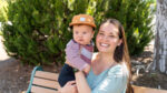 A woman sits on a bench outdoors, smiling while holding a baby wearing a striped shirt and brown cap. Green foliage is in the background.