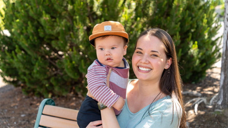 A woman sits on a bench outdoors, smiling at the camera and holding a baby wearing a brown cap and striped shirt. Green trees and sunlight are in the background.