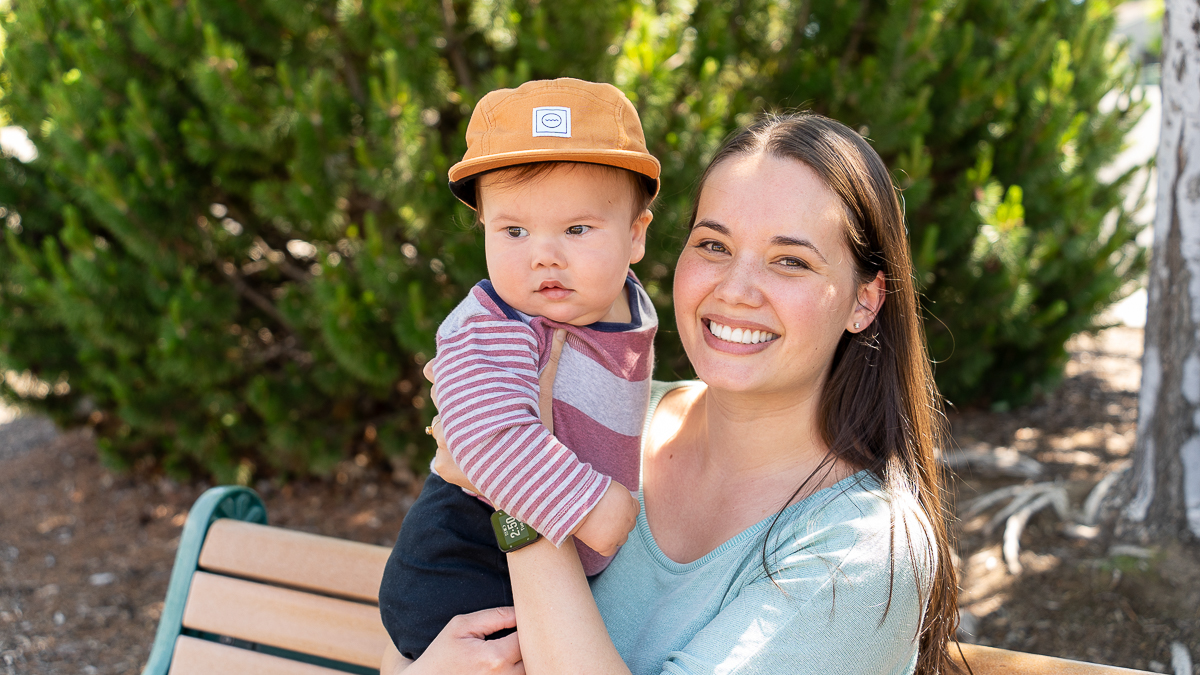 A woman sits on a bench outdoors, smiling while holding a baby wearing a striped shirt and tan cap, with green bushes in the background.