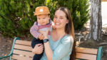 A woman sitting on a bench outdoors holds a baby wearing a striped shirt and brown hat. Both are smiling. Greenery and a tree are visible in the background.