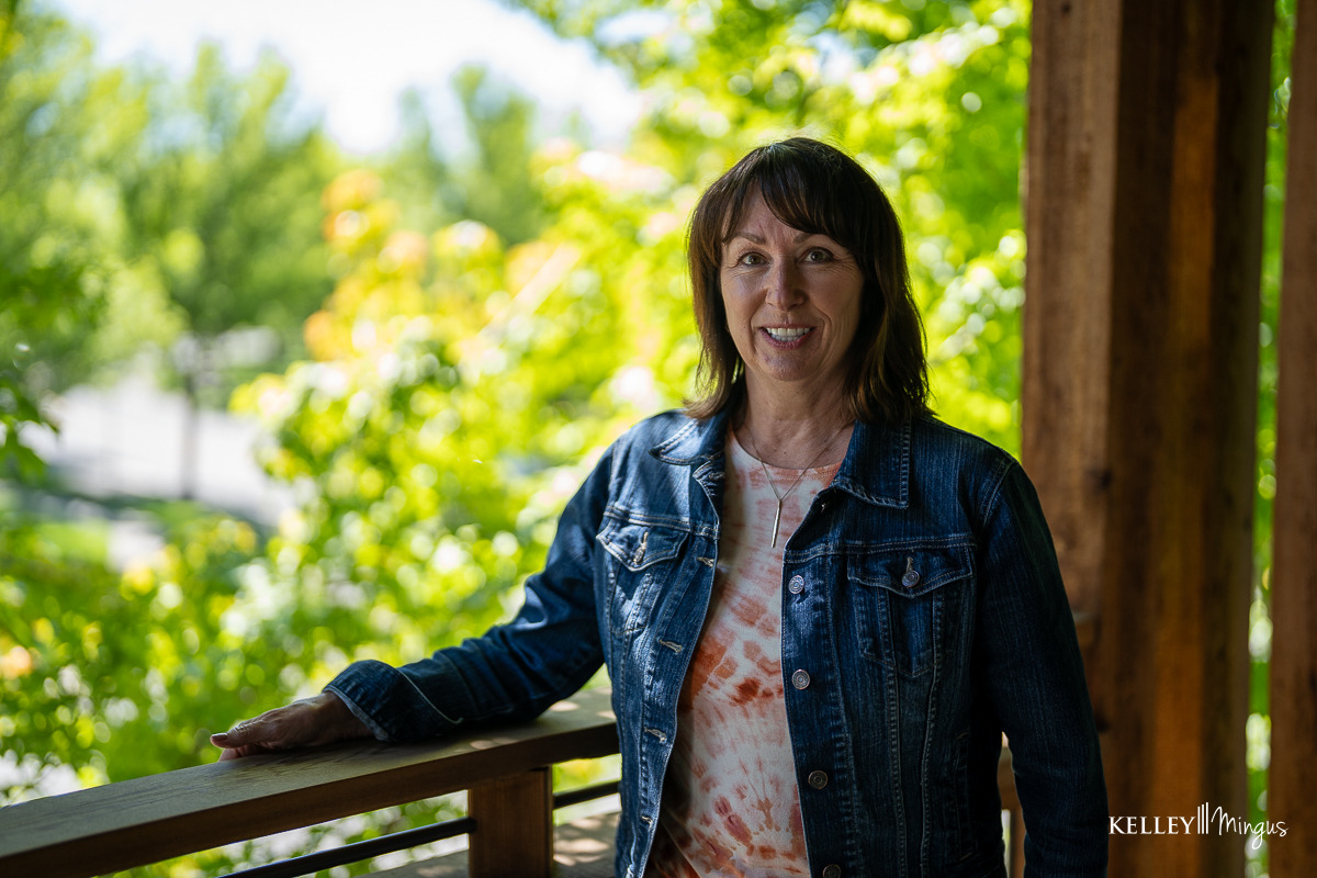 A woman stands on a wooden balcony, wearing a denim jacket and tie-dye shirt, with green trees in the background—thinking about how to choose porcelain veneer shade for her perfect smile.