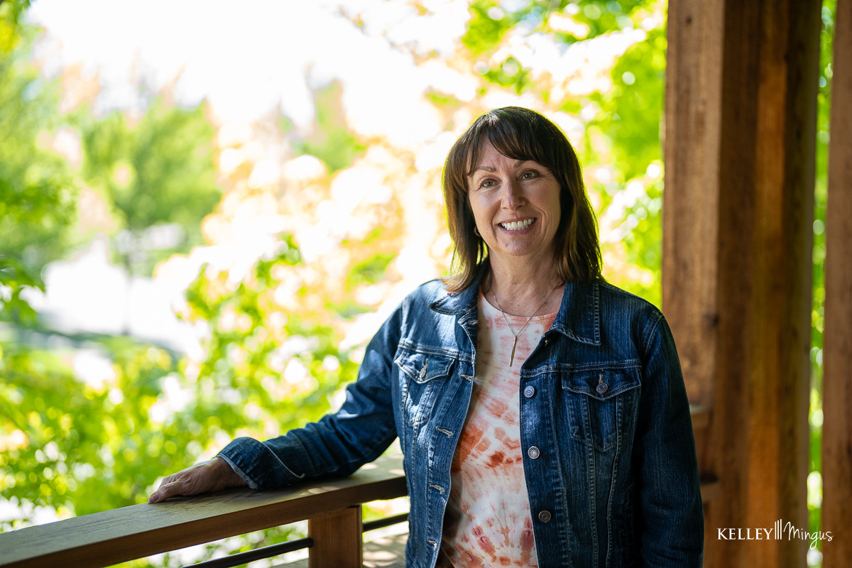 A woman with a bright smile, enhanced by porcelain veneers for gap teeth, stands on a wooden porch in a denim jacket and tie-dye shirt. Green foliage glows in the sunlit background.