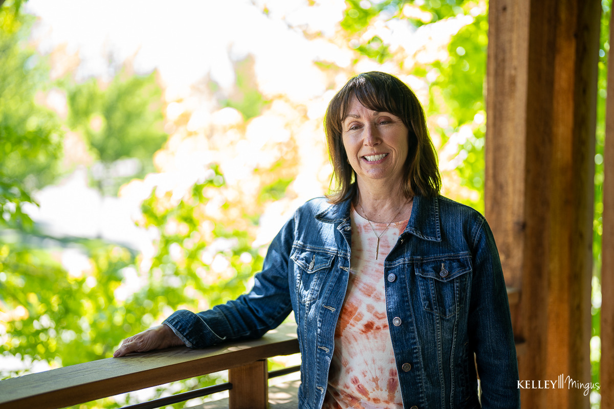 A woman in a denim jacket stands on a wooden balcony, her bright smile—enhanced by custom porcelain veneers—shining in the sunlight with greenery in the background.