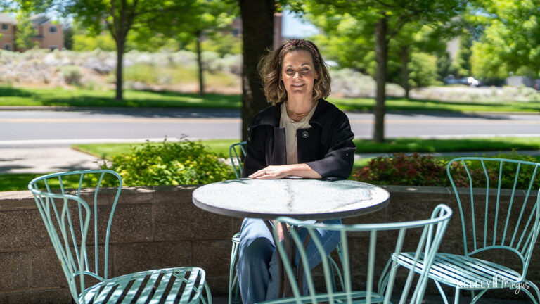 A woman sits alone at a round metal table with empty chairs in an outdoor setting on a sunny day, enjoying relief after her TMJ treatment for migraines.