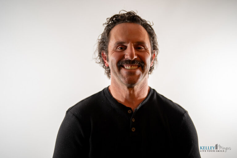 A man with curly hair and a mustache, wearing a black henley shirt, smiles in front of a plain white background. The logo "Kelley Minigus Live Your Smile" is in the corner, reflecting the benefits of holistic TMJ treatment.
