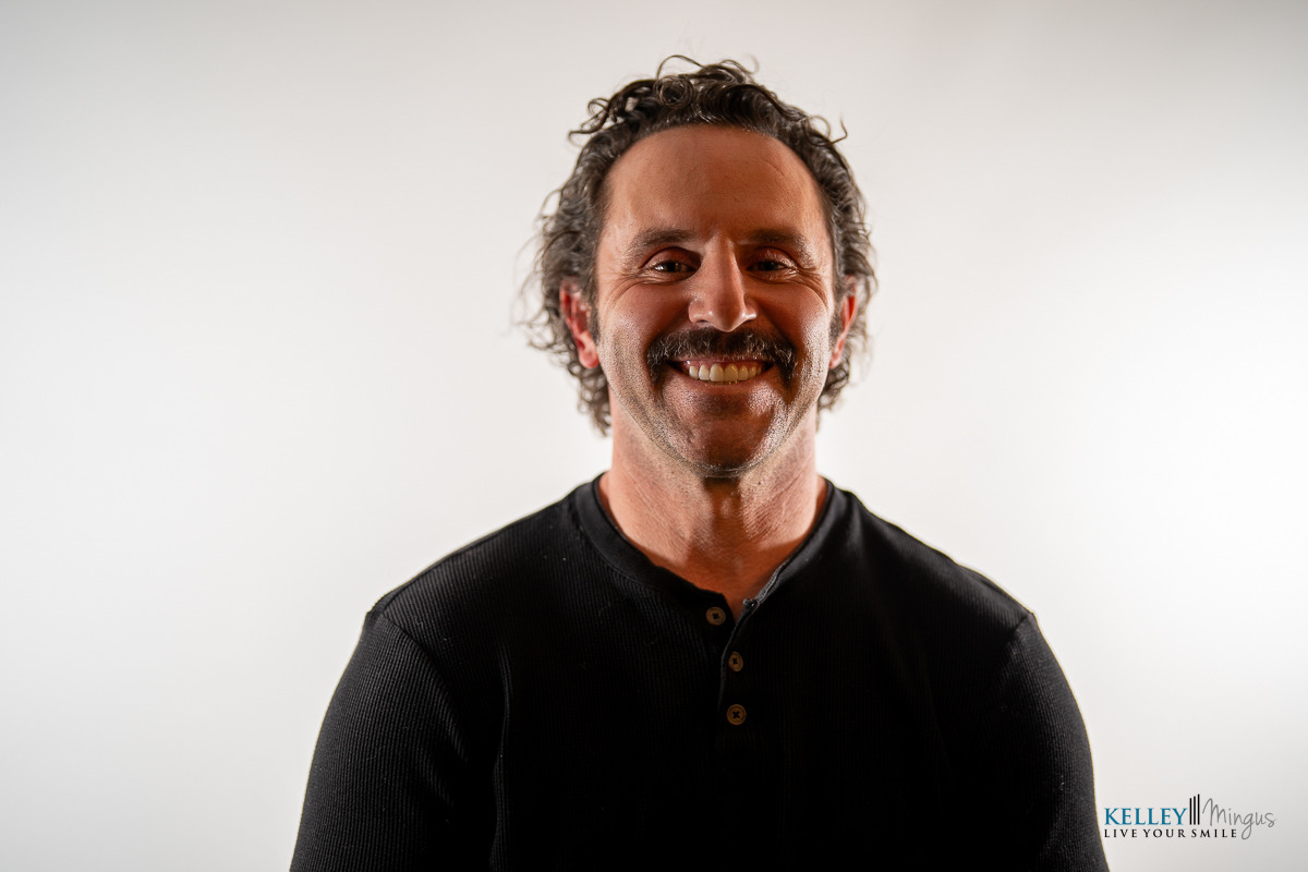 A man with curly hair and a mustache, wearing a black henley shirt, smiles in front of a plain white background. The logo "Kelley Minigus Live Your Smile" is in the corner, reflecting the benefits of holistic TMJ treatment.