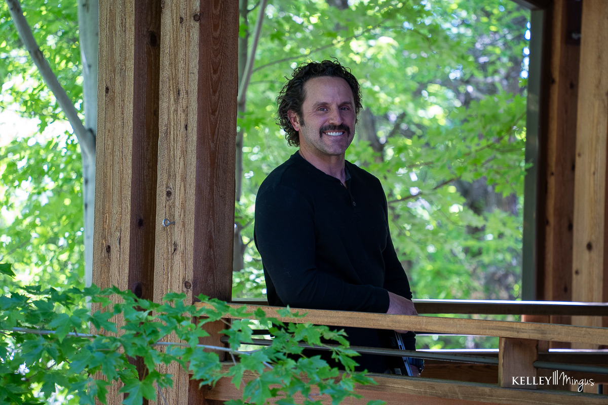 A man with curly hair and a mustache, wearing a black shirt, stands on a wooden porch surrounded by green trees, reflecting on holistic TMJ therapy options.