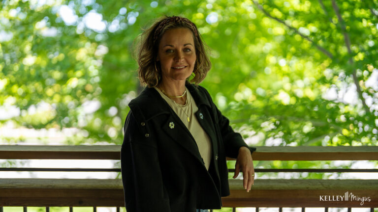 A woman with curly hair stands outside by a wooden railing, wearing a black jacket and light shirt, with green trees in the background—perfectly embodying the natural balance promoted by holistic approaches to dental care.
