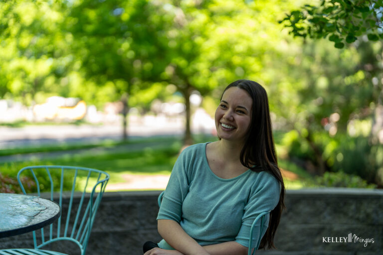 A woman with long brown hair sits and smiles outdoors at a metal table with chairs, surrounded by green trees on a sunny day, reflecting on understanding metal-free restorations.