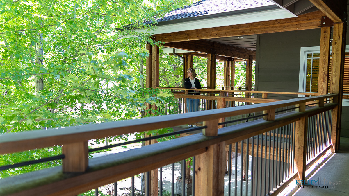 A person stands on a wooden porch surrounded by trees, reflecting on holistic TMJ therapy options while looking out over the railing on a bright day.