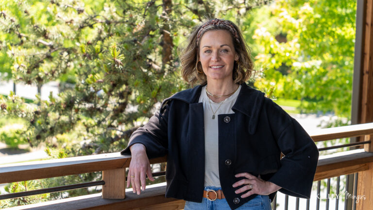 A woman with shoulder-length brown hair stands on a wooden deck, resting one arm on the railing, surrounded by trees and greenery—an ideal setting for embracing holistic TMJ treatment.