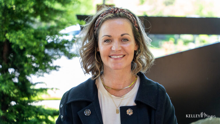 A woman with shoulder-length wavy hair, wearing a headband, gold jewelry, and a black jacket, stands outdoors in front of greenery and a blurred background—perfectly capturing the welcoming atmosphere of general dentistry services in Bend.