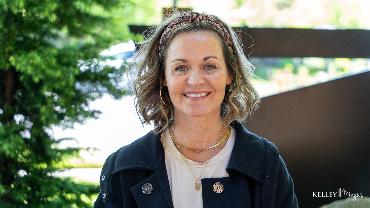 A woman with shoulder-length wavy hair, wearing a headband, gold jewelry, and a black jacket, stands outdoors in front of greenery and a blurred background—perfectly capturing the welcoming atmosphere of general dentistry services in Bend.