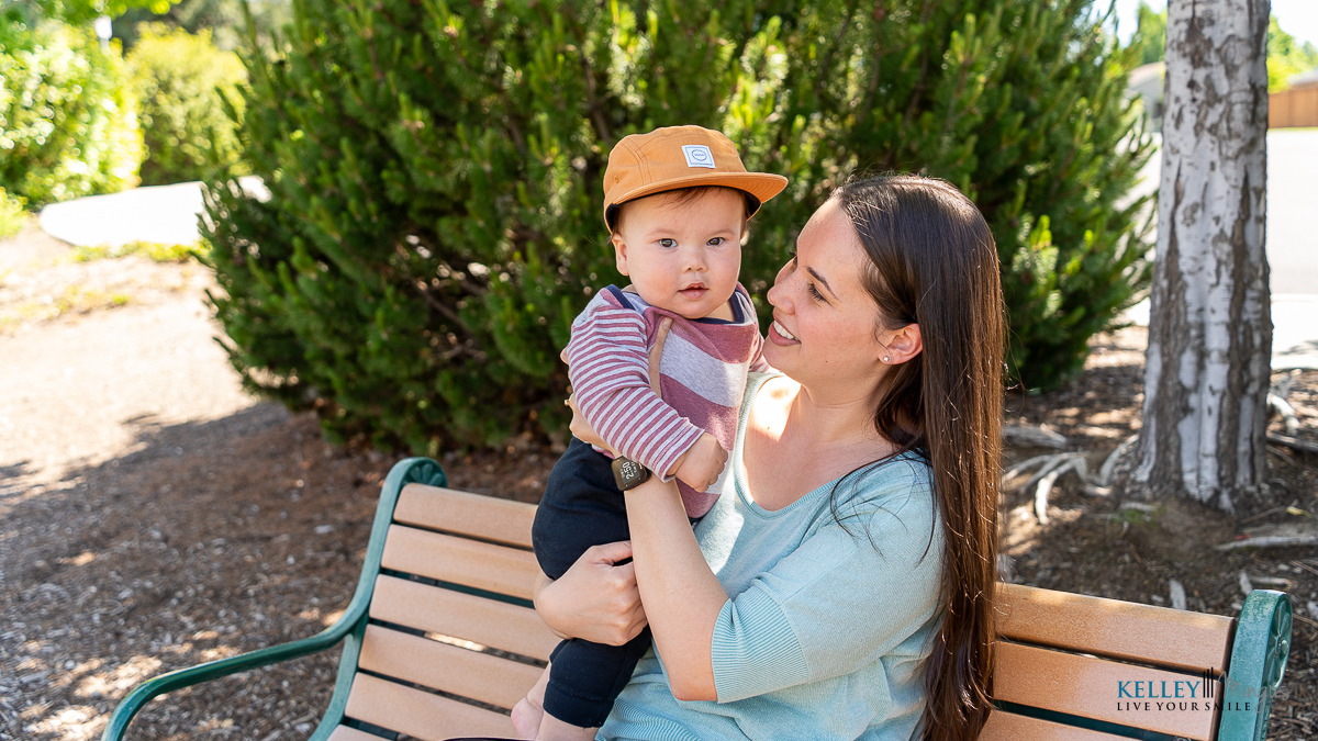 A woman sits on a park bench holding a baby in her lap, the little one in a striped shirt and orange cap. Surrounded by trees, she enjoys peaceful moments—perfect for discussing TMJ treatment for migraines with loved ones.
