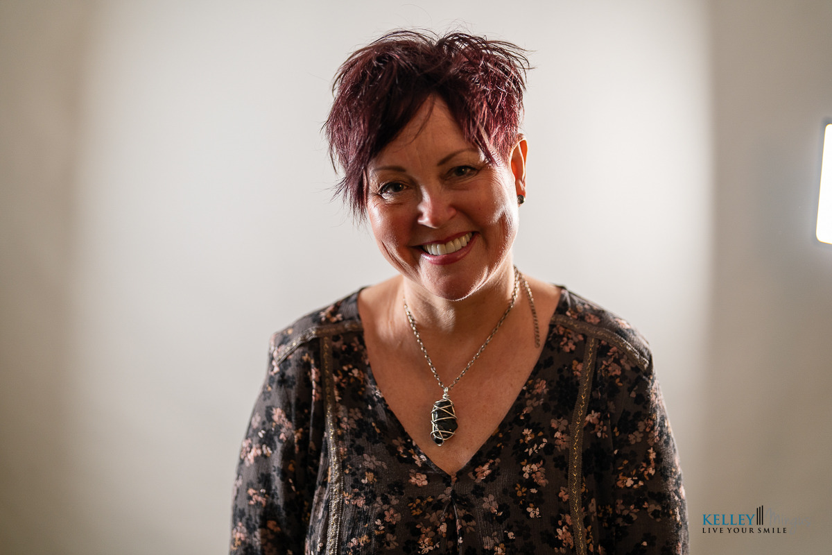 Woman with short dark hair and a patterned blouse smiles at the camera, wearing a large wire-wrapped pendant necklace. The softly lit, plain background highlights her calm presence, reflecting her expertise in holistic TMJ therapy options.