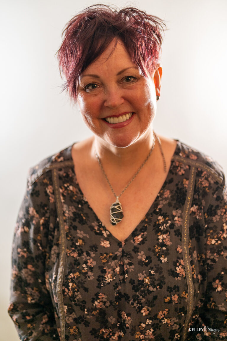 A woman with short, dark red hair smiles at the camera, showcasing the confidence that cosmetic dentistry for busy professionals provides. She wears a floral top and a geometric pendant necklace, standing against a light background.
