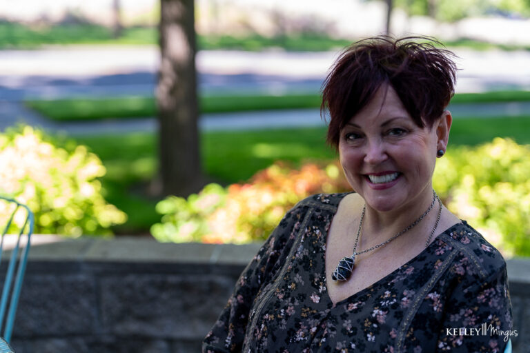A woman with short dark hair smiles outdoors, sitting in front of a stone wall with trees and greenery in the background, radiating confidence after holistic full mouth rehabilitation.