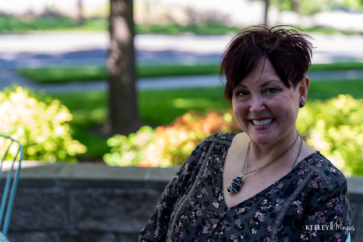 A woman with short dark hair smiles outdoors, sitting in front of a stone wall with trees and greenery in the background, radiating confidence after holistic full mouth rehabilitation.
