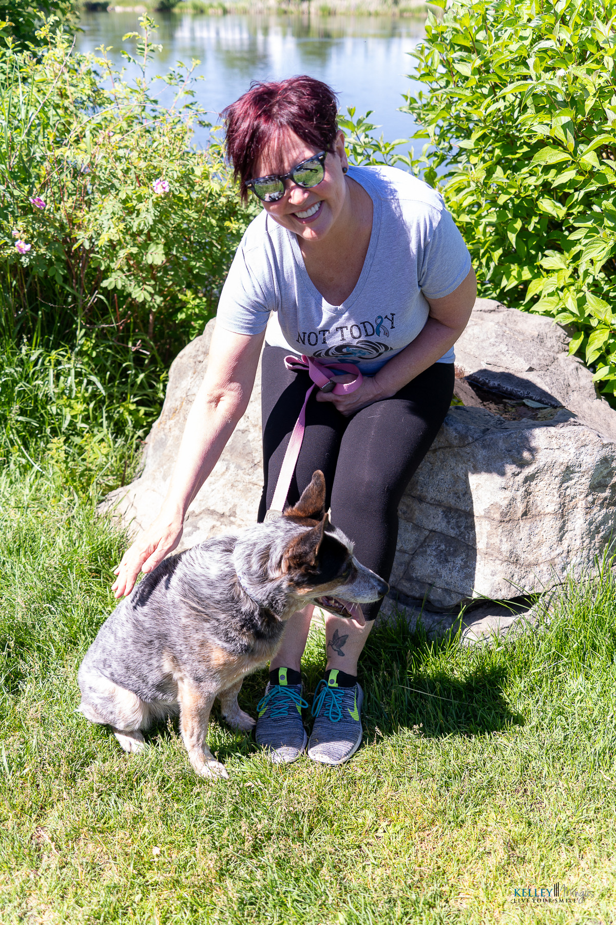Woman in sunglasses sits on a rock by a pond, smiling and showing off her custom porcelain veneers while petting a gray and brown dog on the grassy area during a sunny day.
