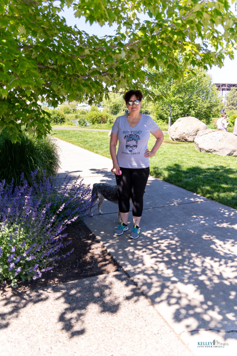 A woman wearing sunglasses and a graphic t-shirt stands on a sunny sidewalk, partially shaded by tree branches, with a small dog beside her and greenery in the background, embodying the calm confidence inspired by holistic TMJ treatment.