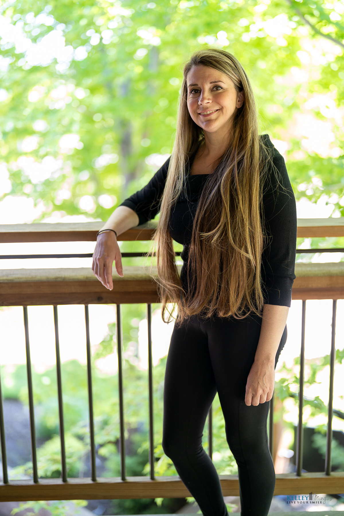 A woman with long brown hair, dressed in black, stands by a wooden railing outdoors, surrounded by trees and greenery—a peaceful scene that reflects holistic approaches to dental care.
