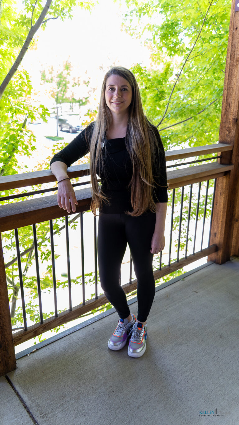 A woman with long brown hair stands on a porch, wearing a black outfit and colorful sneakers. Green trees and a wooden railing are in the background, creating a serene setting that reflects holistic approaches to dental care.